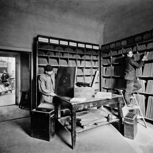 Archivio fotografico d'epoca in bianco e nero: scaffali di scatole fino al soffitto, una persona al tavolo e una su una scala