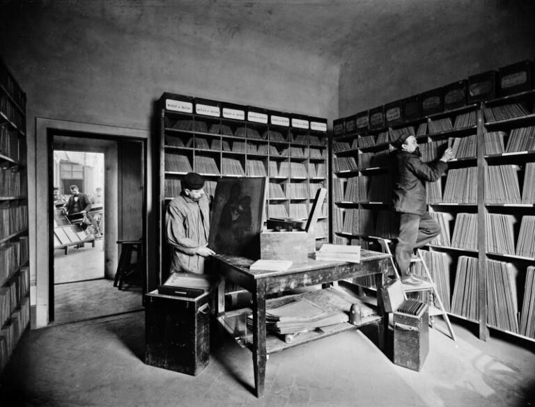Archivio fotografico d'epoca in bianco e nero: scaffali di scatole fino al soffitto, una persona al tavolo e una su una scala
