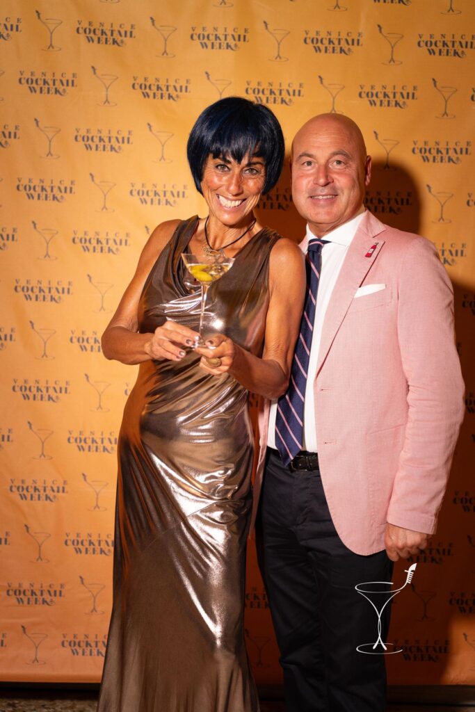 Paola Mencarelli in abito argento, cocktail in mano, e Stefano Santoni in giacca rosa davanti al pannello dell’evento