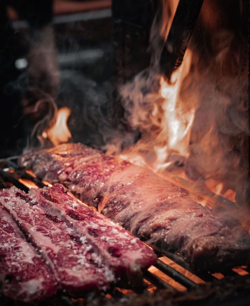 Costillar di manzo sulla griglia della parrilla, tra fiamme e fumo, accanto ad altri tagli di carne ancora crudi