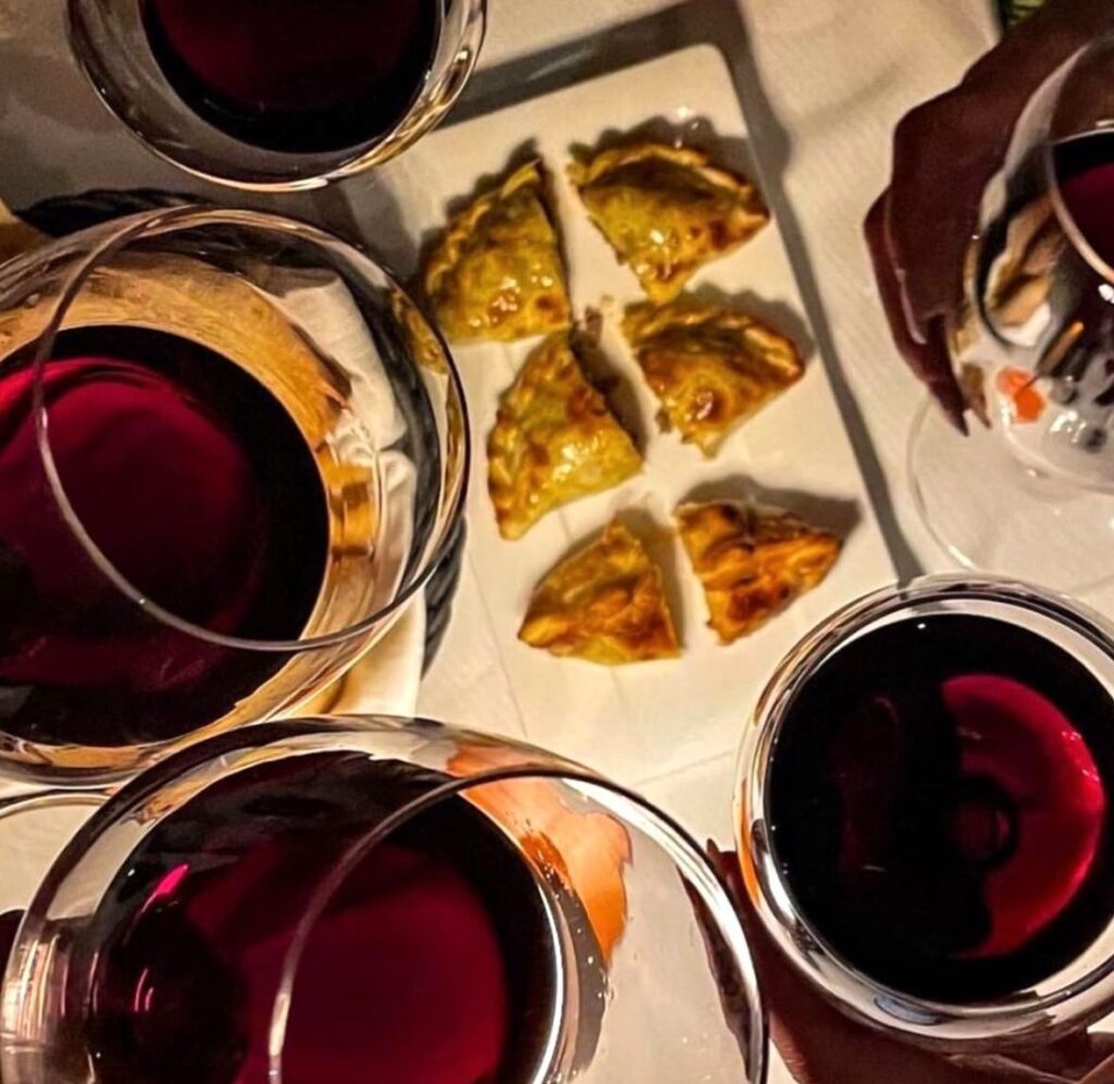 Piccole empanadas su un piatto bianco viste dall’alto, circondate da calici di vino rosso sul tavolo