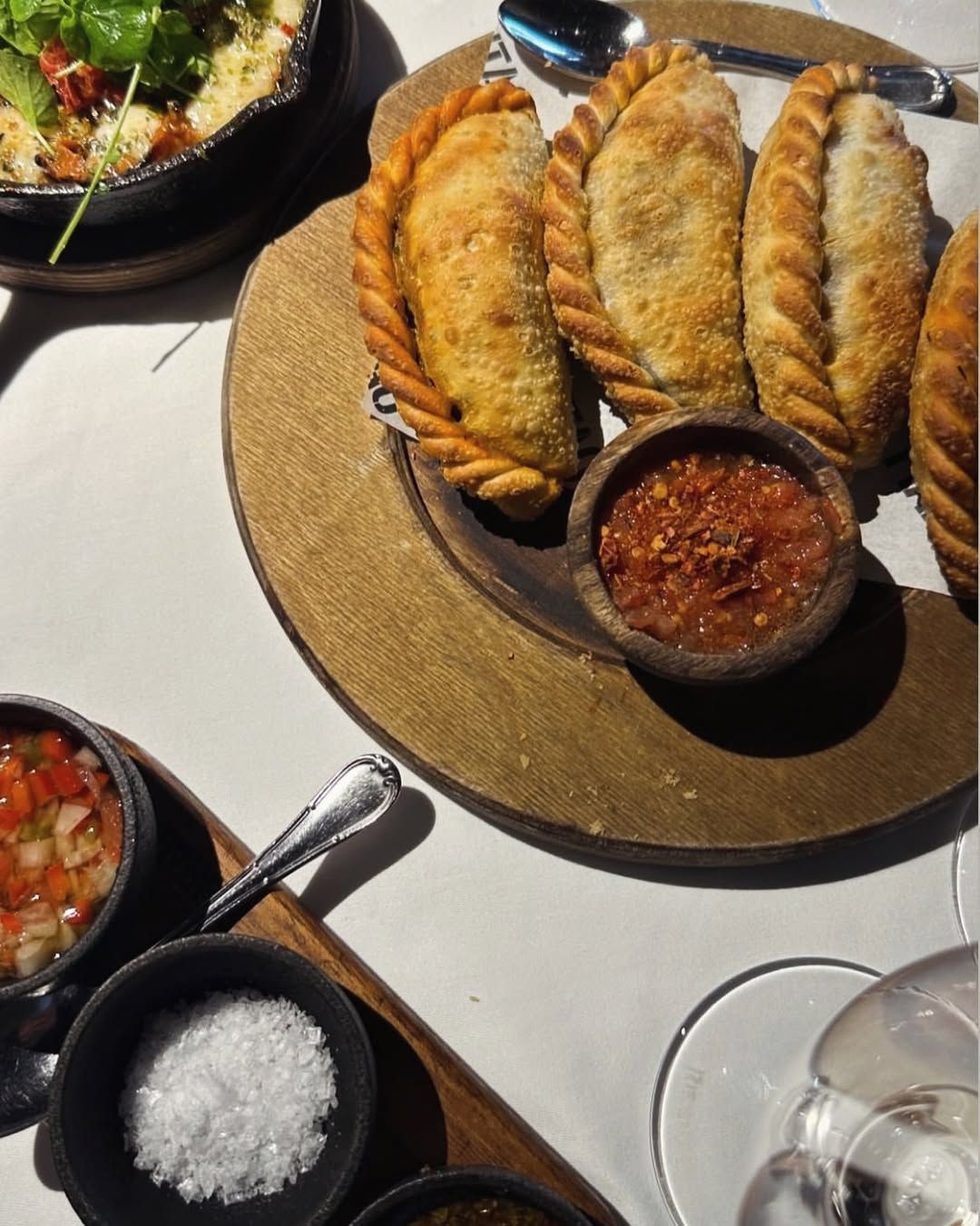 Empanadas al forno su un tagliere di legno, con una ciotola di salsa rossa, sale grosso e altre salse sul tavolo
