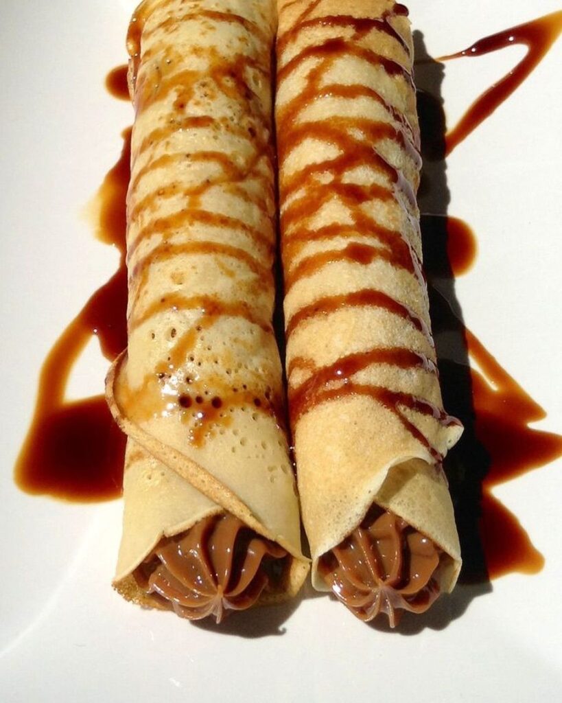 Due panqueques arrotolati e ripieni di dulce de leche, con strisce di salsa scura sopra, su un piatto bianco