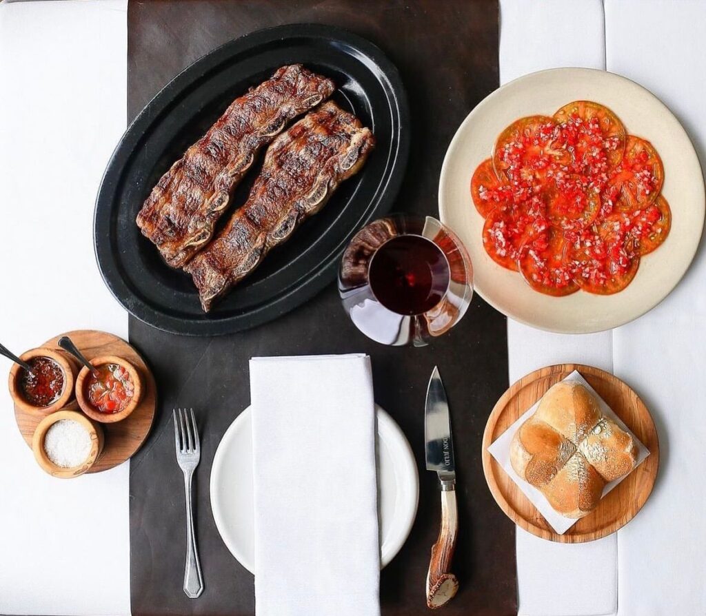 Tavola vista dall’alto: costine alla griglia su un piatto ovale, pomodori a fette, salse, pane e un calice di vino rosso