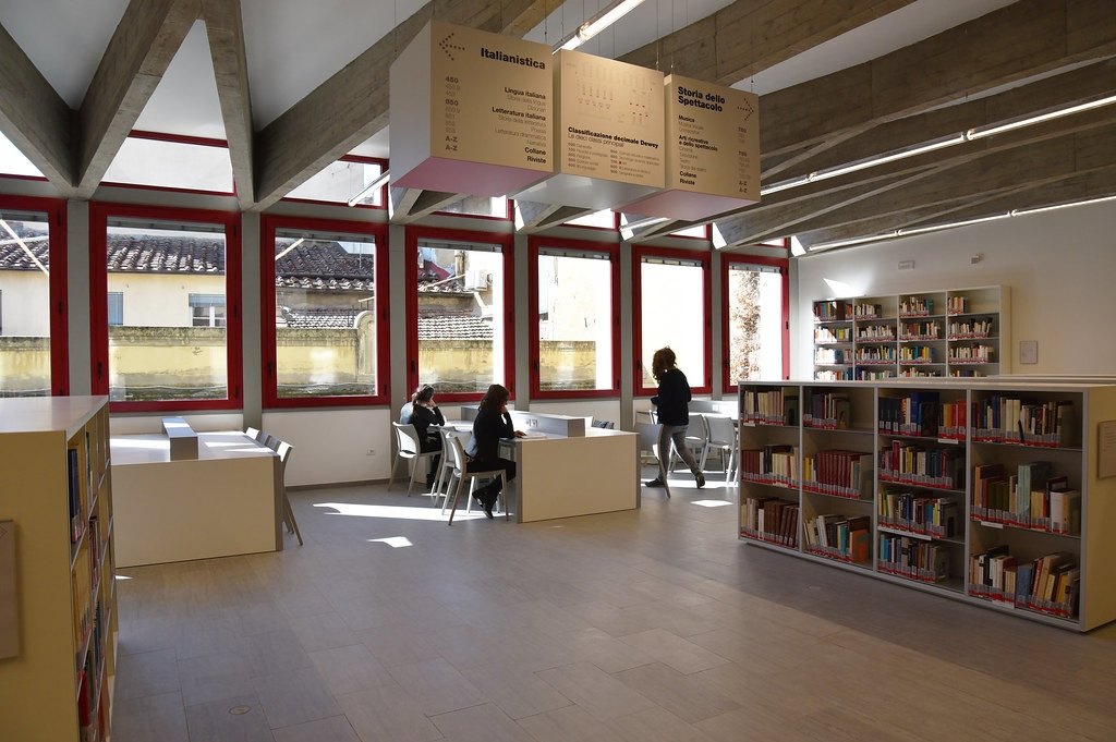 Sala luminosa con scaffali di libri, tavoli bianchi e alcuni studenti al lavoro; grandi finestre con infissi rossi.