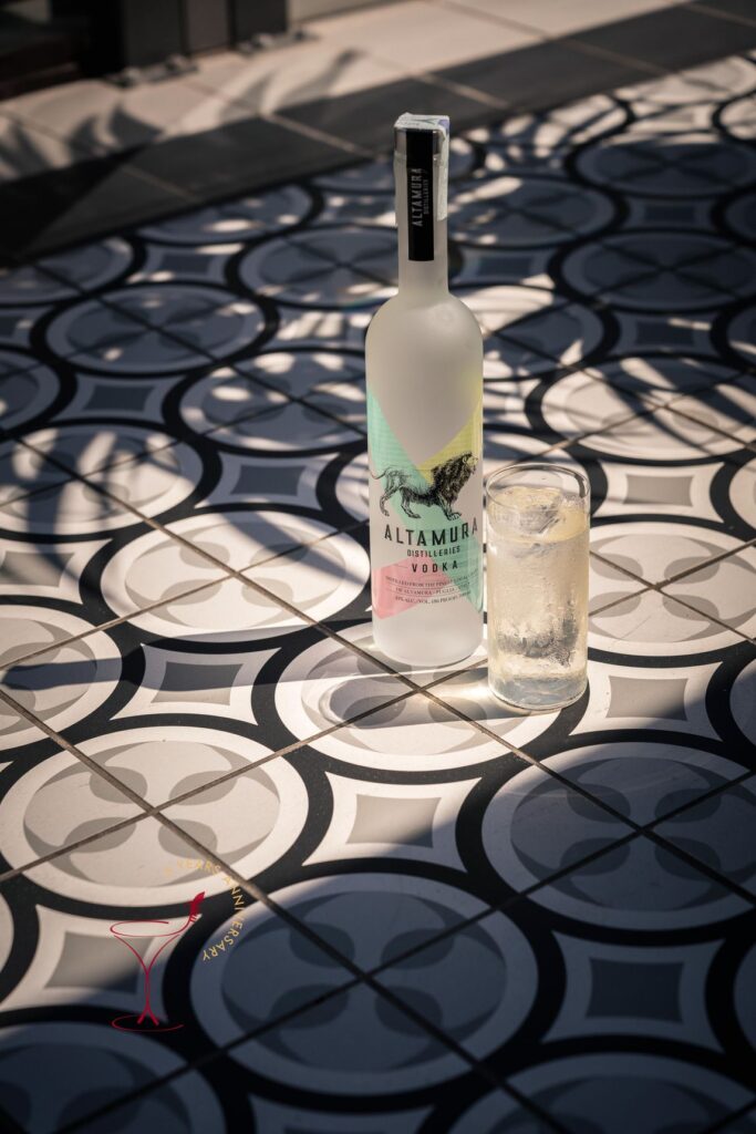 Una bottiglia di vodka Altamura e un drink col ghiaccio su un tavolino a piastrelle bianche e nere, colpito dal sole