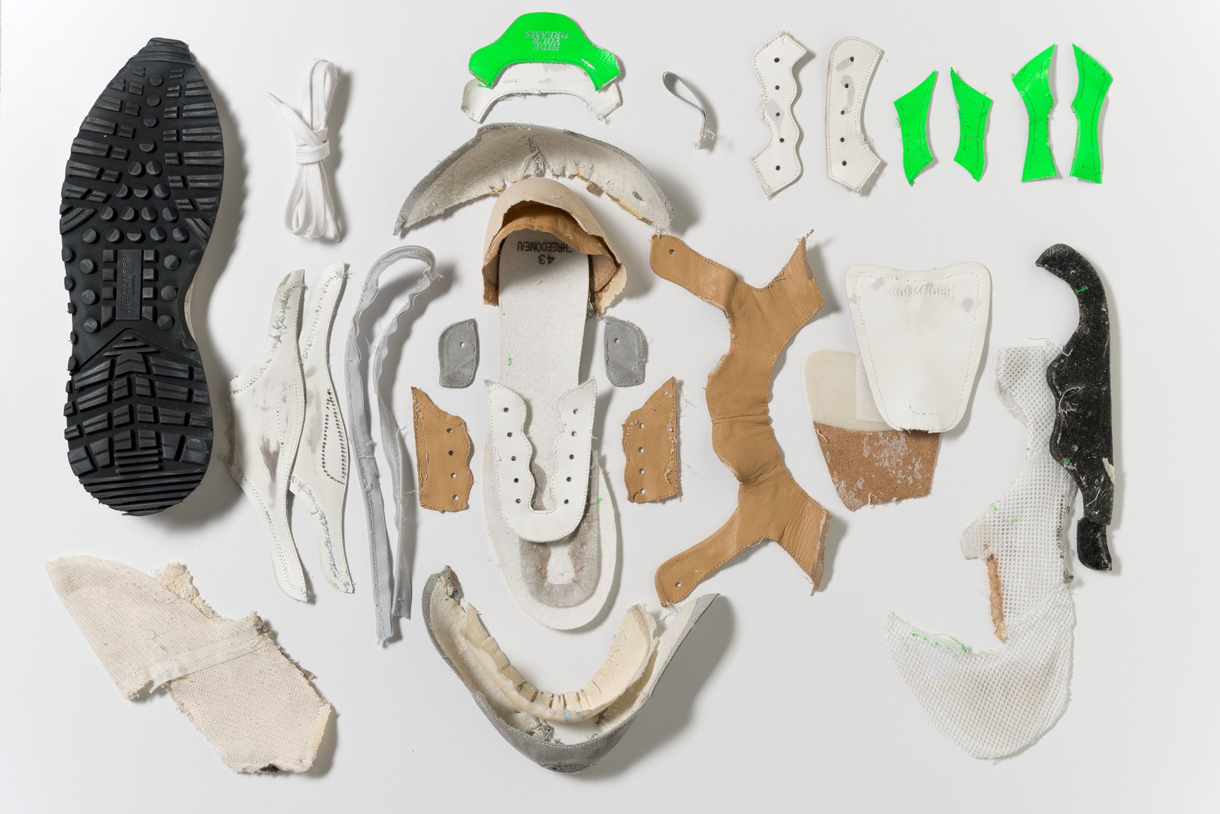 A Hidn-Ander sneaker taken apart piece by piece: sole, laces, upper and green reinforcements laid out in order on a white background