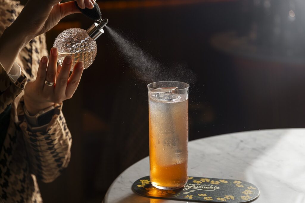 Una mano nebulizza profumo da un flacone di cristallo su un long drink ambrato, posato su un tavolo di marmo