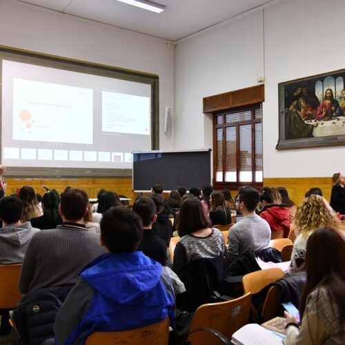 Aula universitaria piena di studenti ripresi di spalle; sul fondo uno schermo con una proiezione e un grande dipinto.