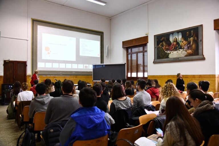 Aula universitaria piena di studenti ripresi di spalle; sul fondo uno schermo con una proiezione e un grande dipinto.