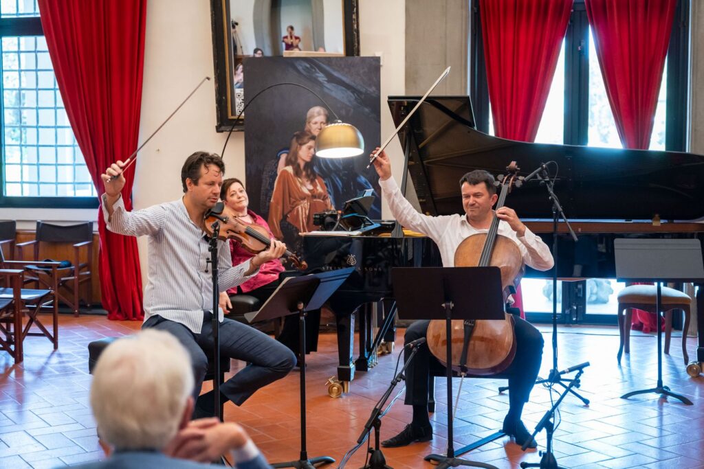 Un violinista e un violoncellista suonano davanti al pubblico in una sala con tende rosse, con il pianoforte alle spalle